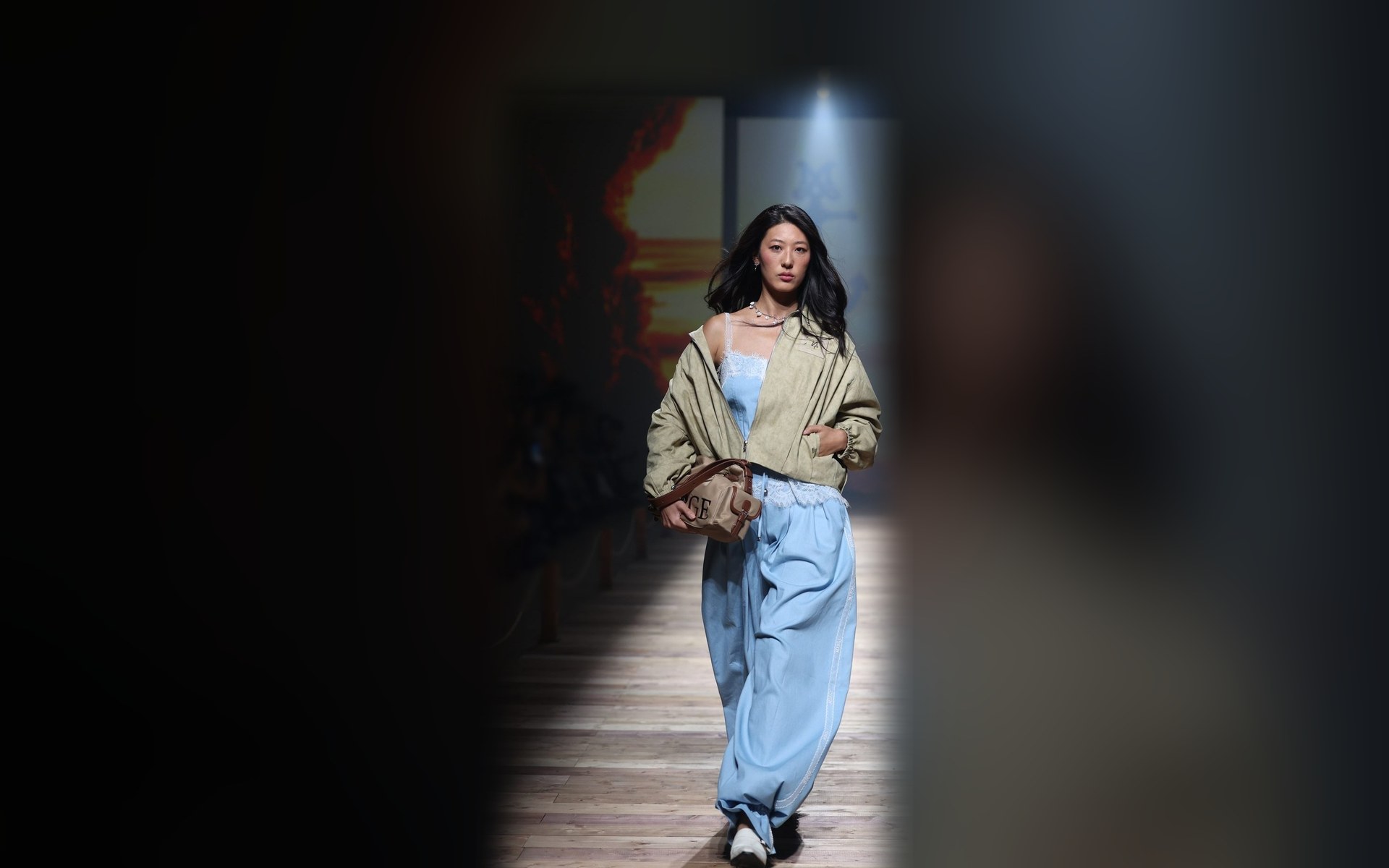 A model walks the MERGE TROPICAL BLISS show at BIFW 2026 in an olive jacket, light blue denim camisole and wide pale blue trousers, carrying a MERGE bag