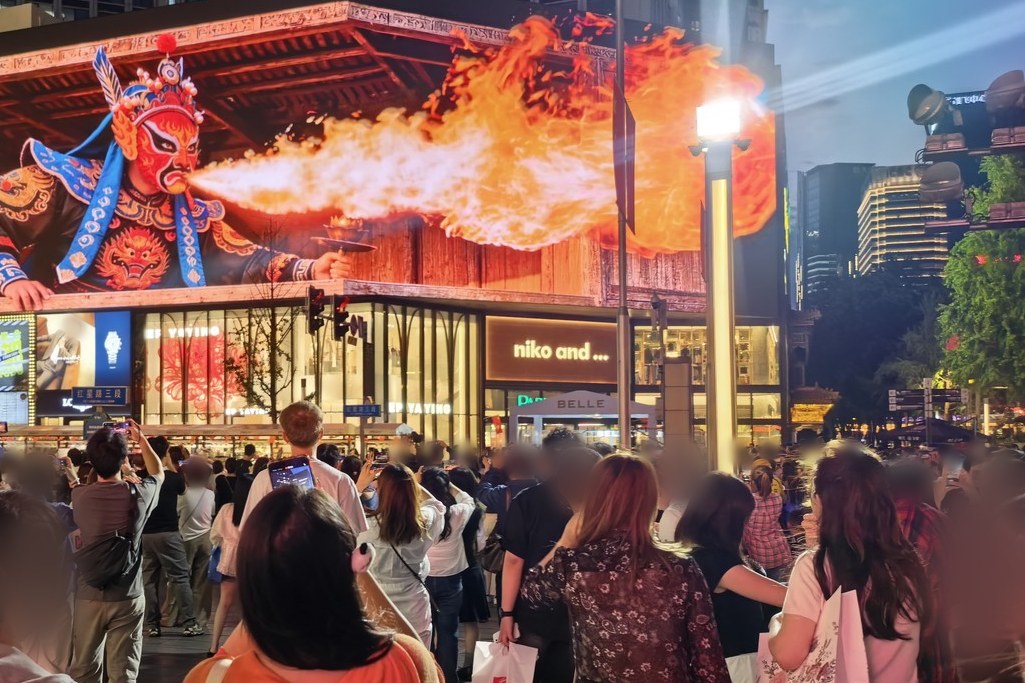 Crowds on Chunxi Road film the naked-eye 3D billboard on Chengdu Times Parkson as a Sichuan opera performer breathes fire; faces are blurred